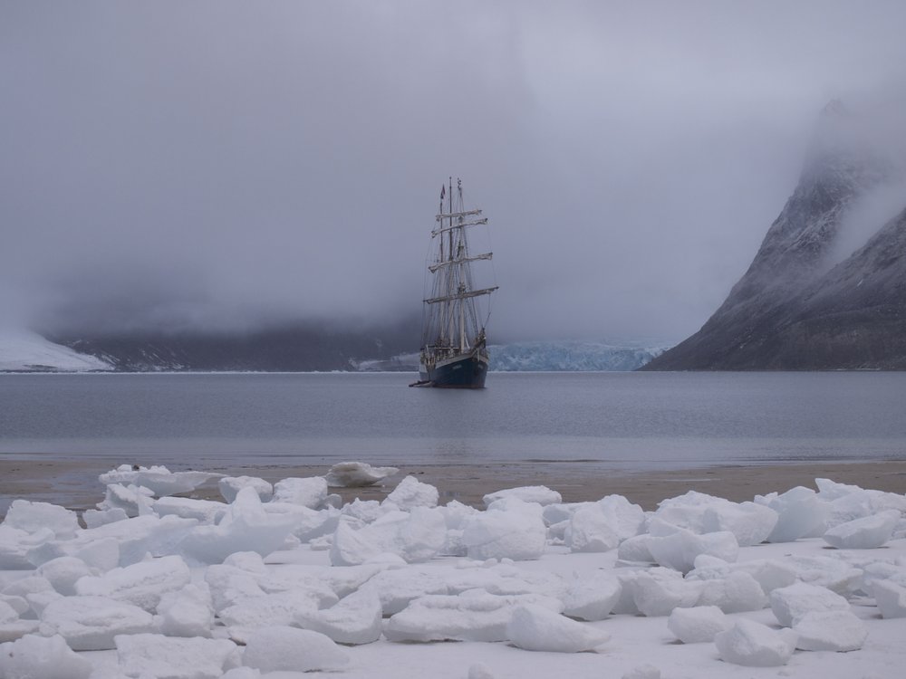 A ship floats in a still, foggy bay that is surrounded by cliffs and chunks of snow. The sails are missing, leaving the mast of the ship a skeleton.